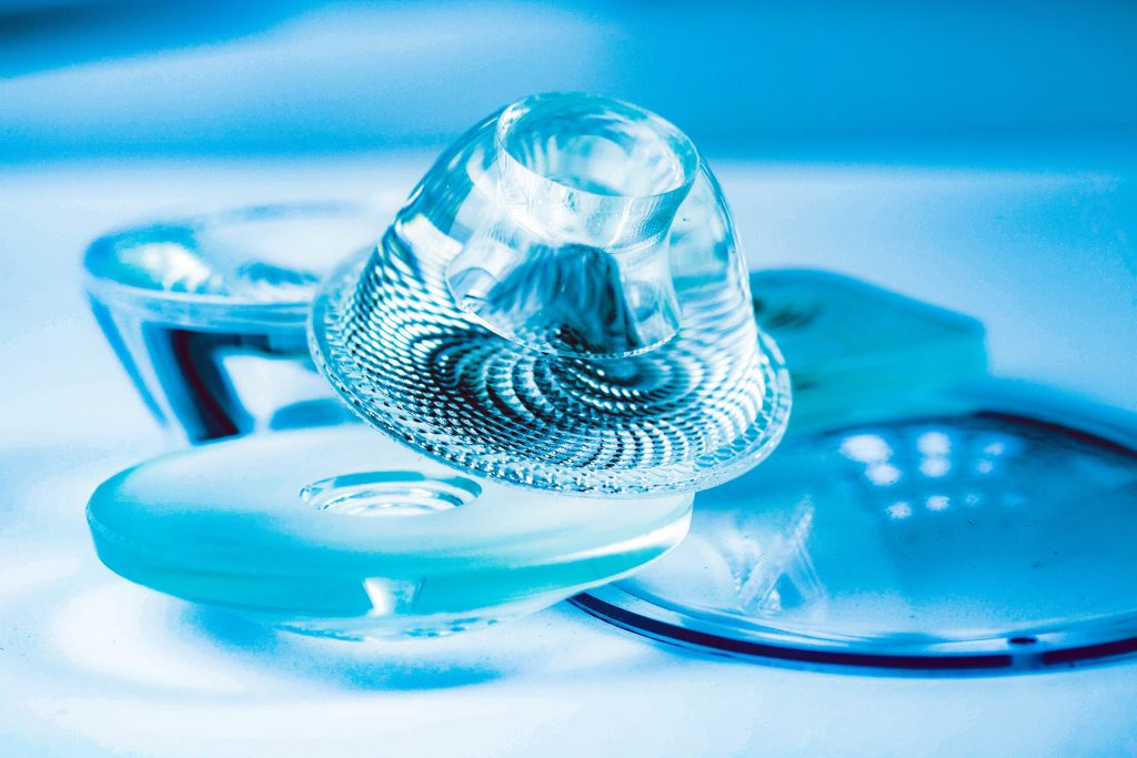 A close-up photograph of various optical components, including lenses and prisms, arranged on a table. The components exhibit different shapes, sizes, and reflective properties, indicating a diverse range of optical elements used in various applications such as imaging systems or scientific instruments.