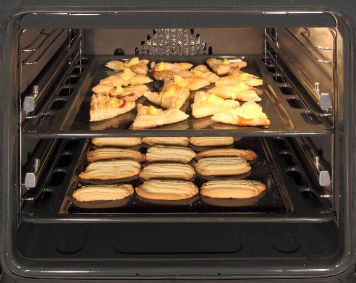 A close-up photograph of freshly baked bread inside a white goods oven. The oven features a glass door, allowing a view of the bread as it bakes. The image conveys the concept of baking in a domestic kitchen environment using household appliances.