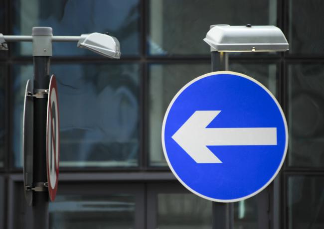 A close-up photograph of a one-way traffic sign on a highway. The sign indicates traffic flow in one direction. The image conveys the concept of traffic regulation and road signage.